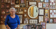 An elderly woman with a beautiful smile, Barbara White, stands in front of a wall in her home covered with a lifetime of photos.